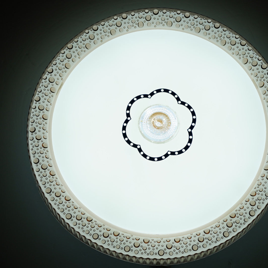 A round ceiling light with a decorative beige-golden floral border and a small black floral motif at the center, flush-mounted on a white ceiling; it emits bright, diffused white light and suits traditional or classic interiors.