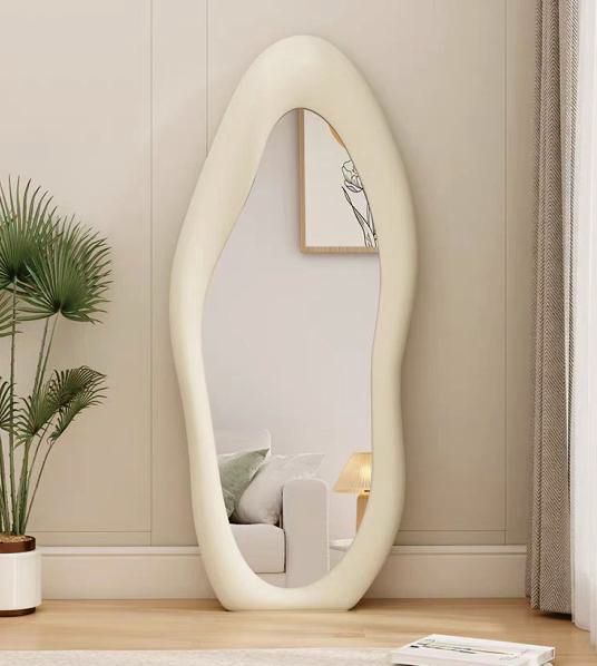 Egg-shaped mirror on a wooden floor with a plant and framed picture in the background.