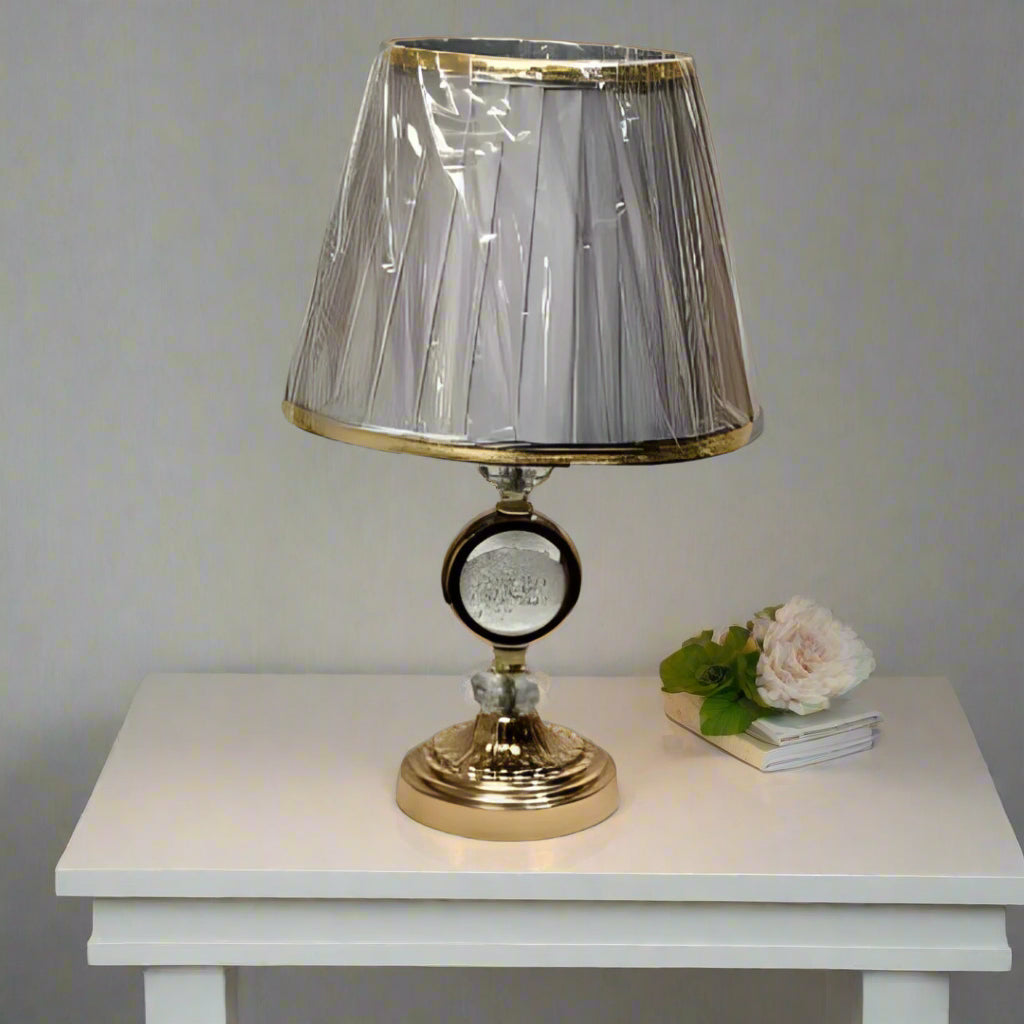 Round gold table lamp with a pleated grey shade with gold trim, featuring a crystal centrepiece, placed on a white table.