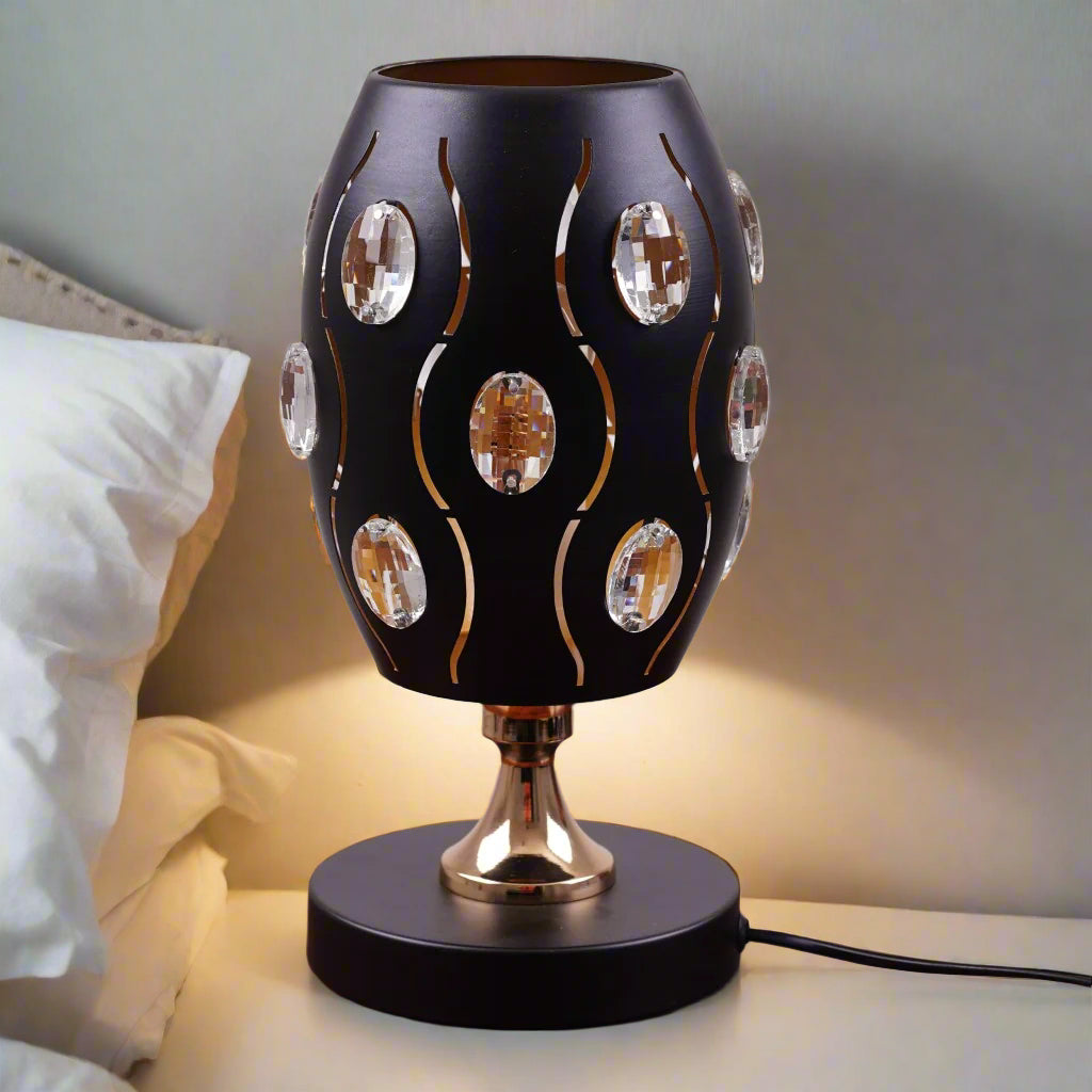 A black globe table lamp with a geometric design, adorned with crystal beads, gold accents, and a sleek base, sit on a wooden table. It features an LED light with an E27 bulb, casting a warm, stylish glow.