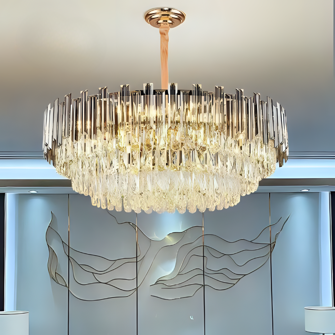 A round crystal chandelier with gold accents hanging from the ceiling in a modern interior, with a decorative wall featuring abstract metallic artwork in the background.