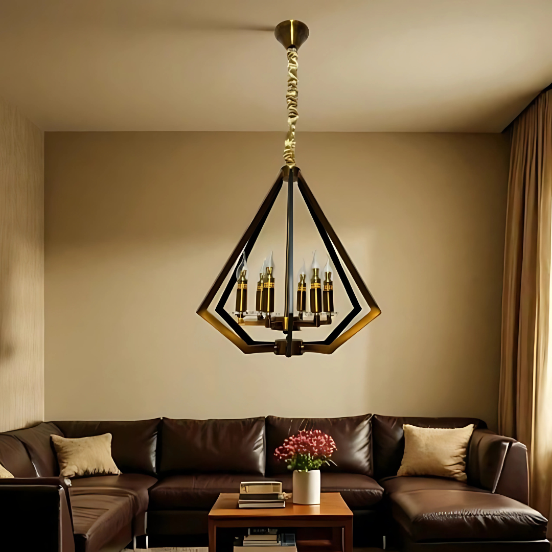 A modern living room with a dark brown L-shaped leather sofa, a wooden coffee table topped with books and a vase of flowers, and a geometric pendant chandelier in the center featuring gold-toned accents and candle-style bulbs. The room has beige walls, floor-length curtains, and a warm-toned wooden floor with a soft area rug.
