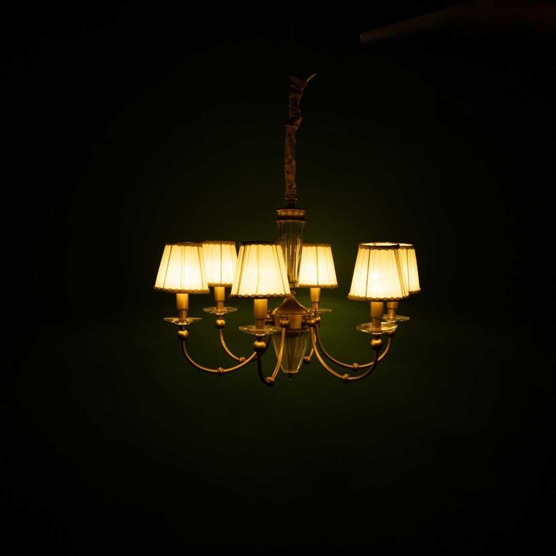 "A golden chandelier, softly illuminated in a dimly lit room, features multiple gracefully curved arms. Each arm holds a white pleated lampshade with intricate decorative trim. At the centre of the chandelier is a beautiful glass column, and it is suspended from a gold-colored rod, adding a touch of elegance to the room's atmosphere.