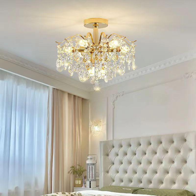 A gold-tone crystal chandelier mounted close to the ceiling in a luxurious bedroom. The fixture has hanging teardrop crystals and gold branches, casting a warm glow over a tufted headboard and elegant curtain backdrop.