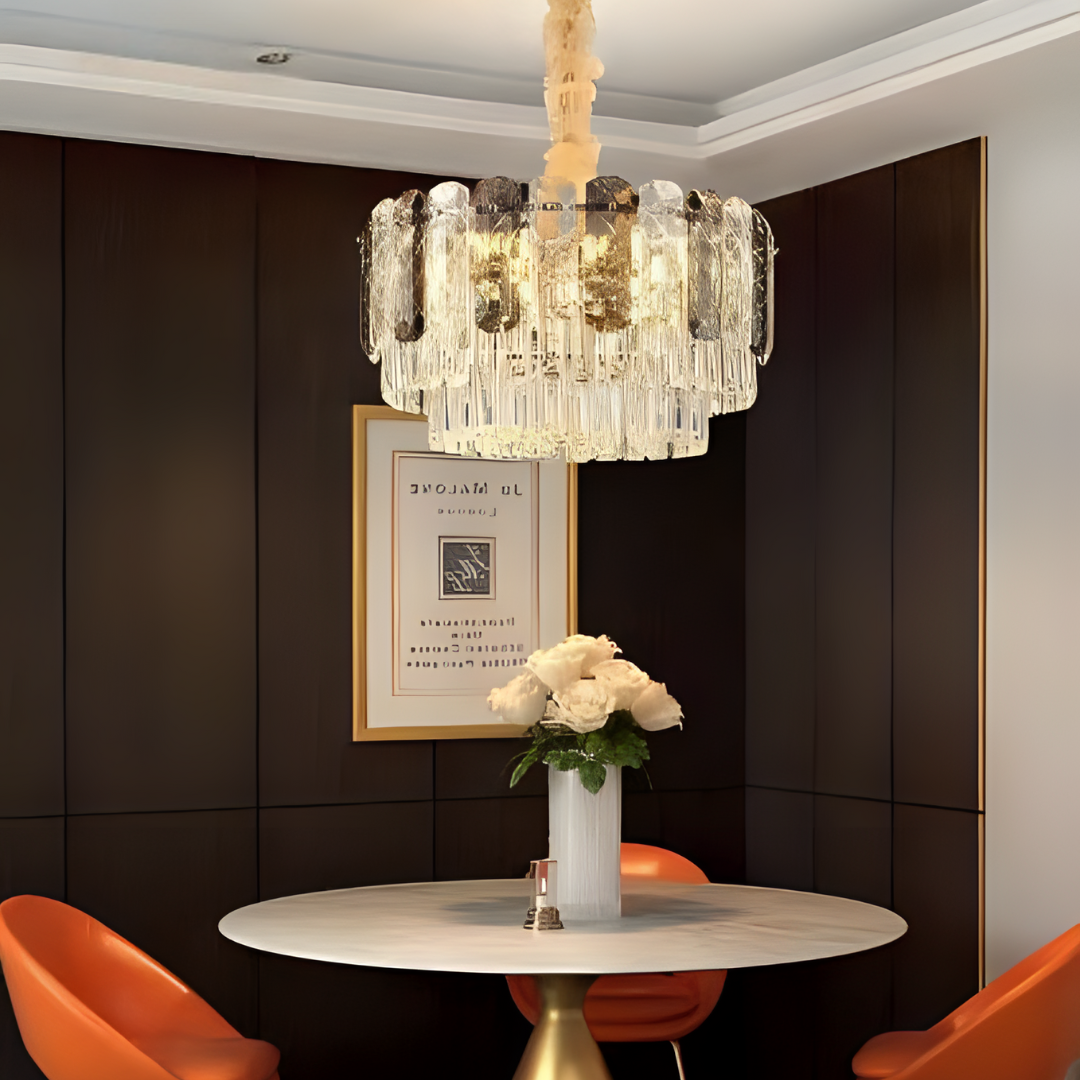 A luxurious crystal chandelier with a gold frame hanging above a round dining table with orange chairs, set against a dark accent wall.
