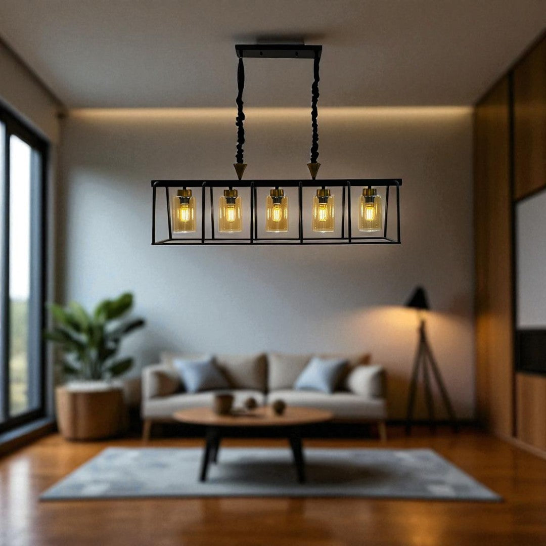 A modern rectangular chandelier with five exposed vintage-style light bulbs enclosed in glass shades, suspended by two twisted black rods. The fixture hangs above a cozy, minimalist living room featuring a beige sectional sofa, a neutral area rug, wooden flooring, and a potted plant in the corner.
