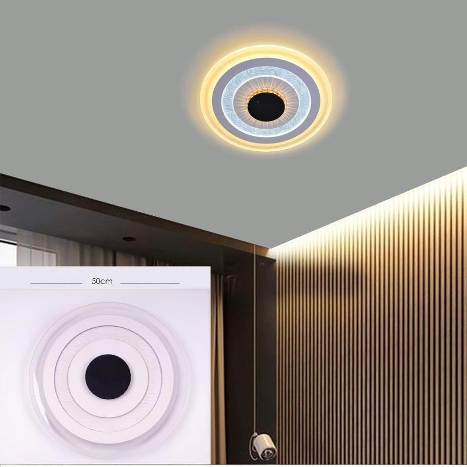 A circular ceiling light featuring concentric LED rings in warm yellow and white with a black center, installed on a grey ceiling corner above vertical wooden wall panels; it emits layered, ambient lighting.
