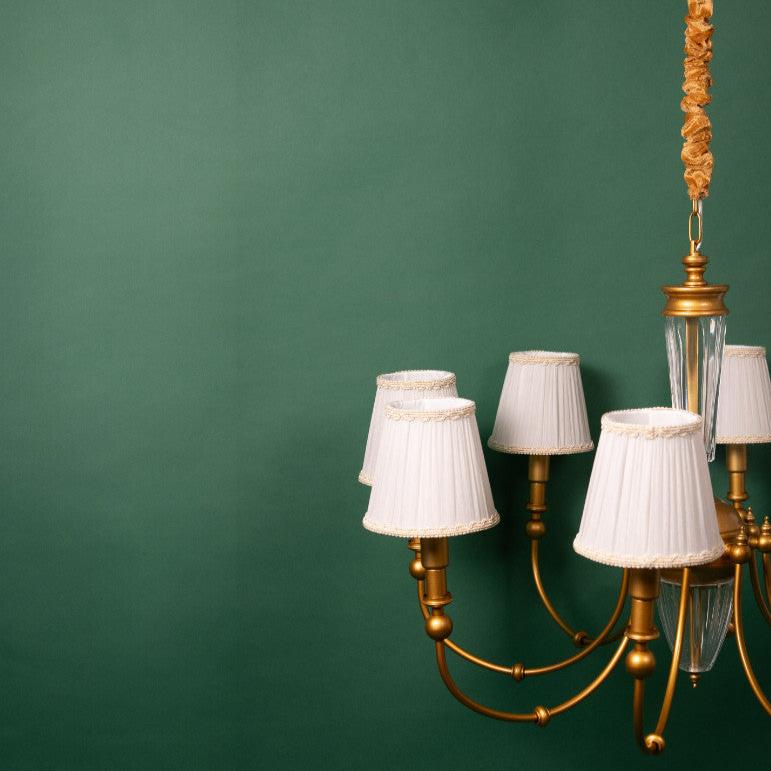 A close-up of a golden chandelier with multiple curved arms, each holding a white pleated lampshade adorned with decorative trim. The chandelier features a central glass column and is suspended from a gold-colored rod.