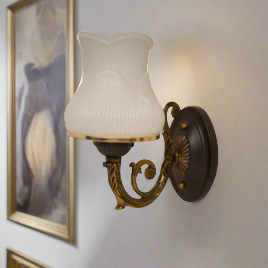 A classic bronze wall sconce with a decorative backplate and a fluted frosted glass shade, mounted on a white wall near framed artwork.