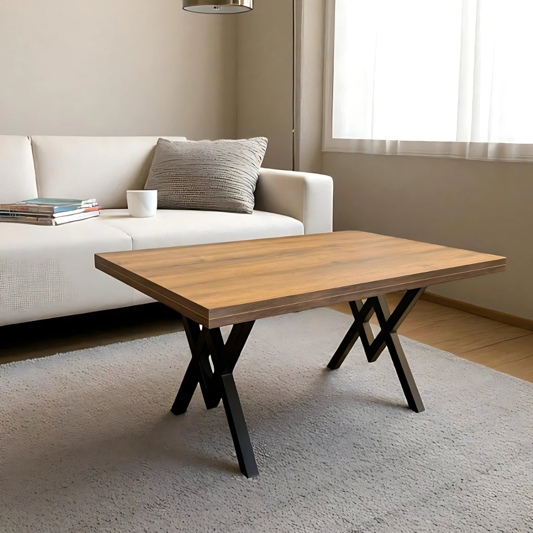 A modern living room featuring a minimalist wooden coffee table with a rectangular top and black crisscross metal legs. The table sits on a light grey rug in front of a white sofa adorned with a textured grey cushion. A stack of books and a white mug rest on the sofa, with soft natural light streaming in from a window with white curtains.
