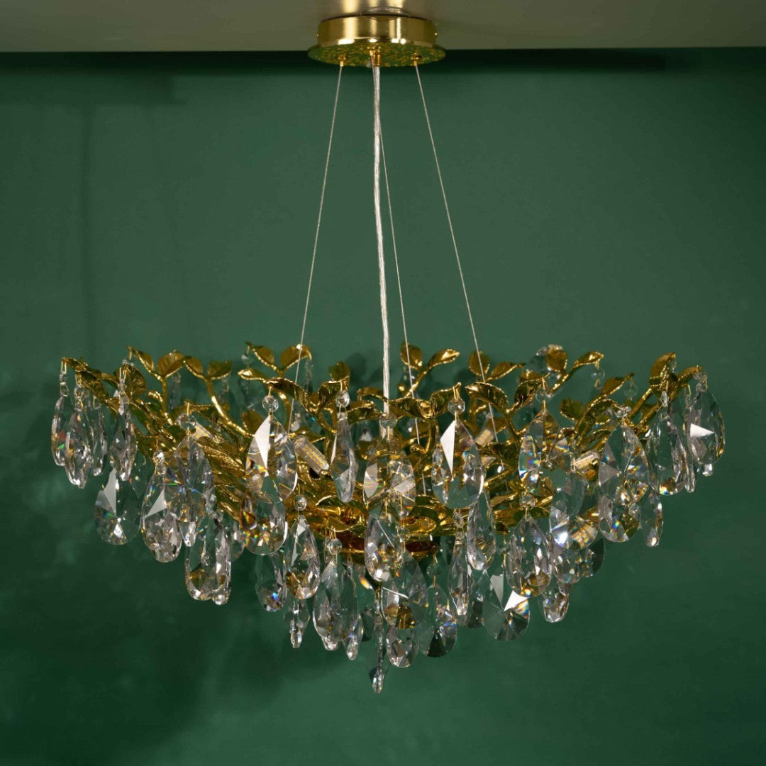 A golden chandelier with a leaf-shaped design and crystal accents, its gold base fixed to a white ceiling, set against a green background.