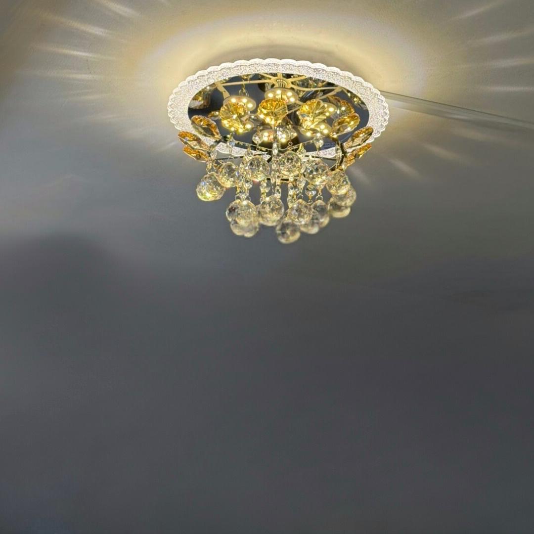 A compact, ceiling-mounted chandelier featuring a gold-toned metal frame and a cluster of teardrop-shaped crystal pendants. The fixture casts warm LED lighting, creating a decorative sunburst reflection on the ceiling.
