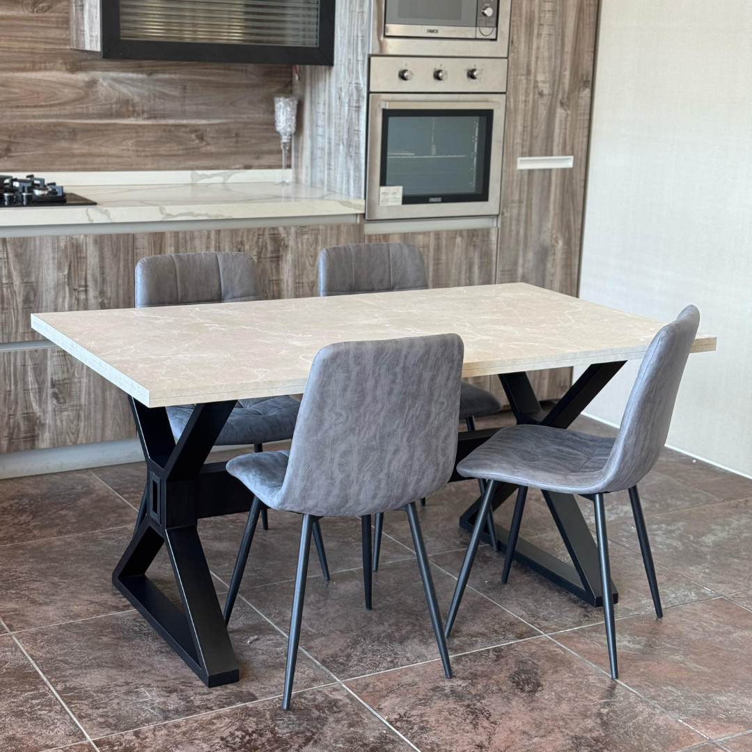 A modern dining set featuring a rectangular wooden-top table with a black metal X-shaped base, paired with four grey faux-leather chairs with metal legs. The set is placed in a contemporary kitchen with wood-paneled walls, built-in stainless steel appliances, and a white countertop