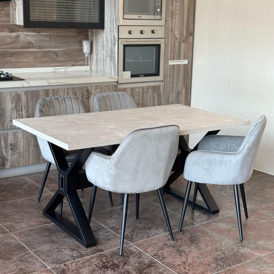 A modern dining set featuring a rectangular wooden-top table with a black metal X-shaped base, paired with four grey chairs with metal legs. The set is placed in a contemporary kitchen with wood-paneled walls, built-in stainless steel appliances, and a white countertop