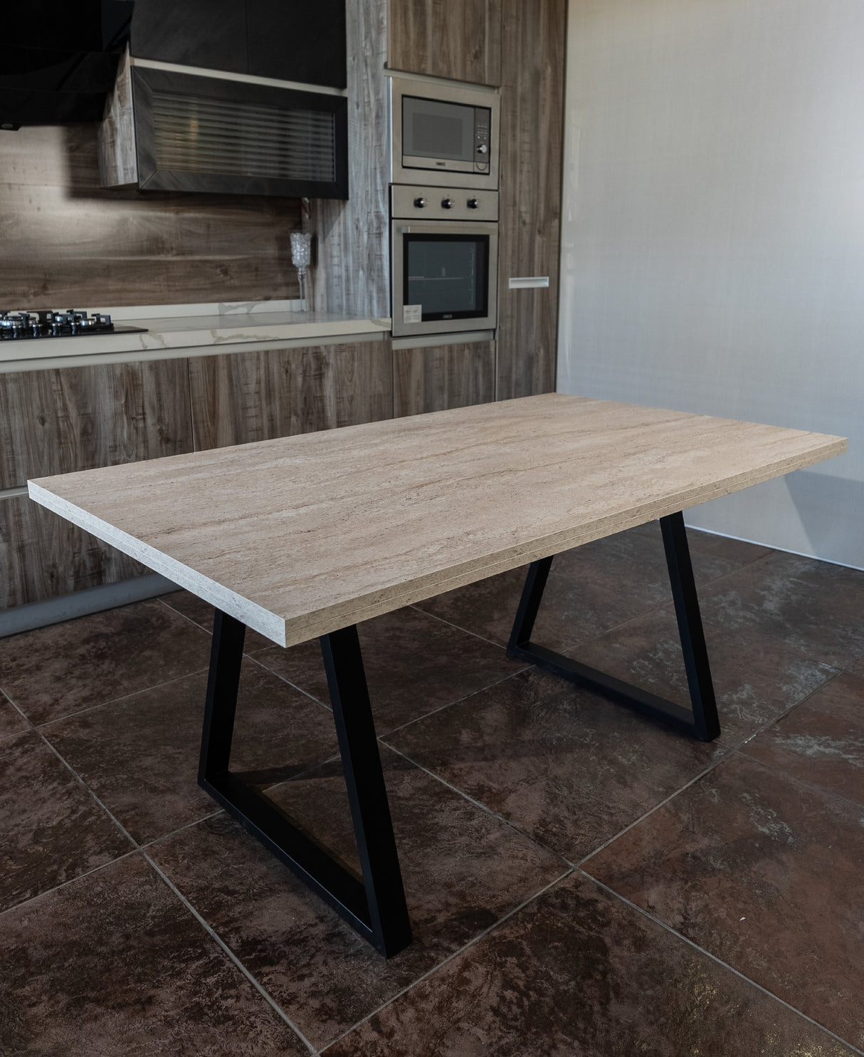 Full view of a modern dining table with a melamine wood top and black steel legs, placed in a contemporary kitchen with brown tiled flooring and built-in stainless steel appliances.