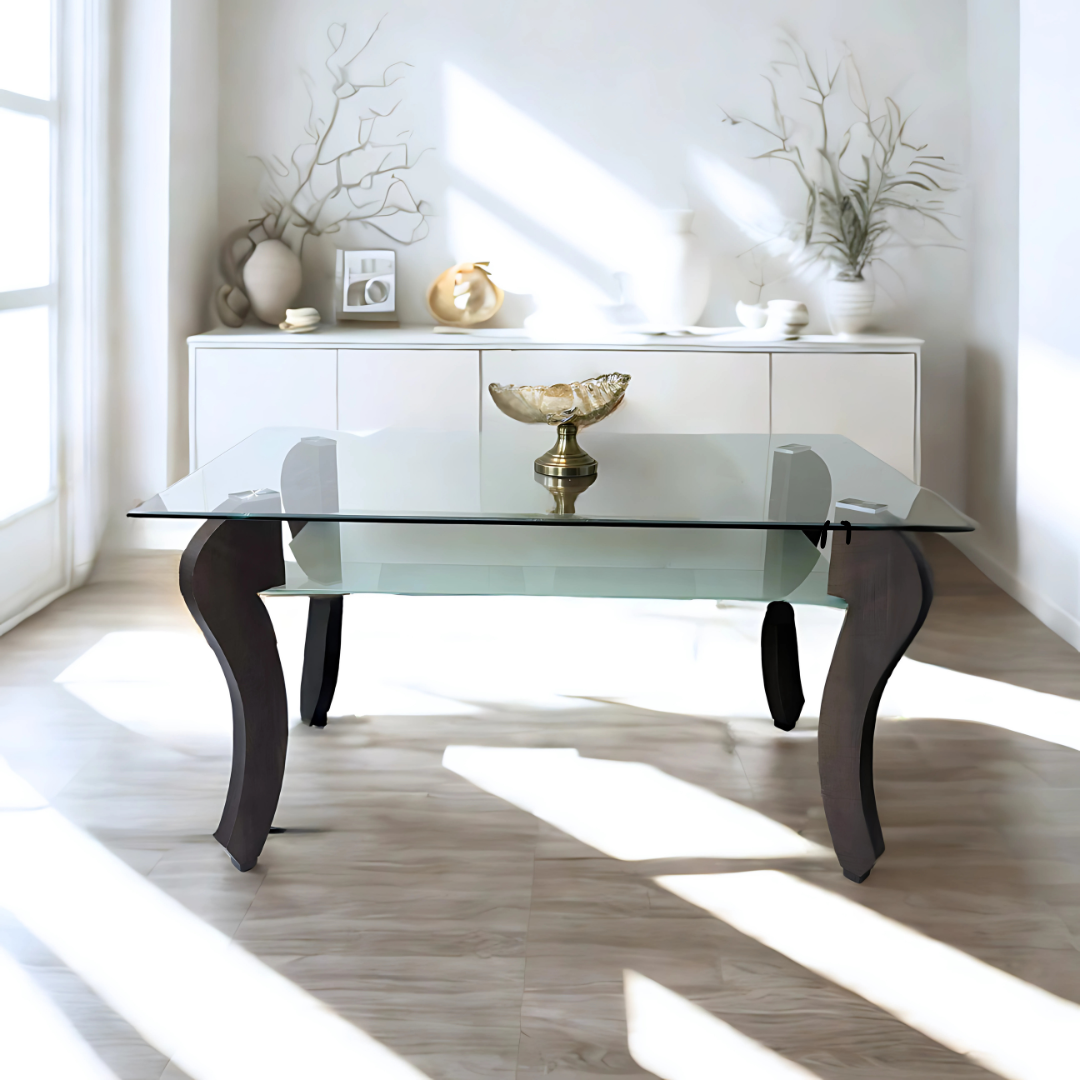 tylish glass-top table with curved dark wooden legs, placed in a bright, minimalist room with light wood flooring and natural sunlight streaming through large windows. A decorative gold bowl sits at the center of the table, and a modern white sideboard with artistic vases and sculptures stands in the background.