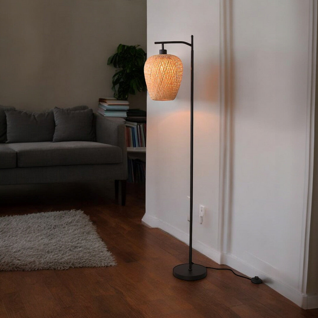 A modern floor lamp with a black stand and a woven lampshade, placed in a cozy living room with a grey sofa and bookshelf in the background.