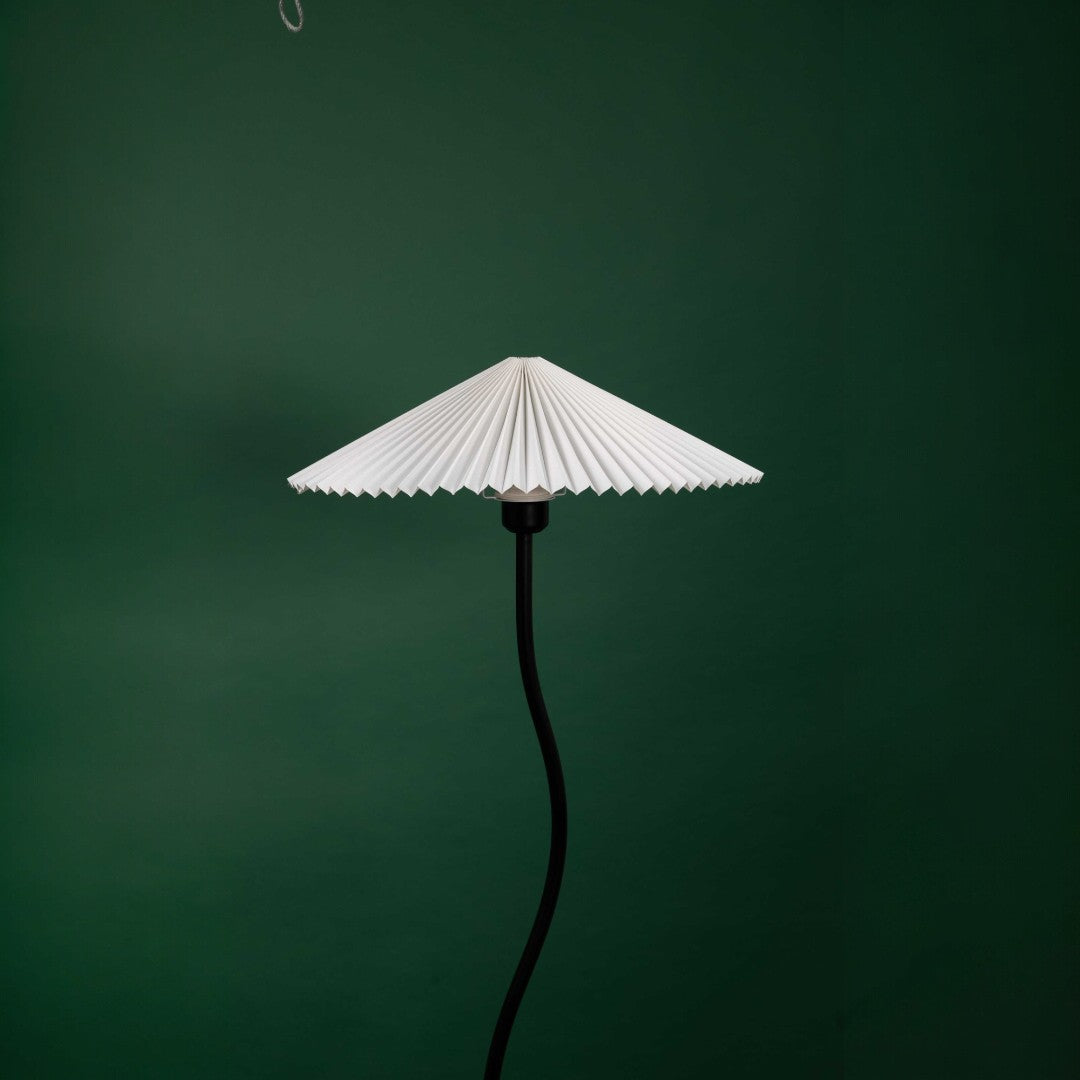 A black floor lamp with a curved stand and a pleated white lampshade, photographed against a green backdrop.