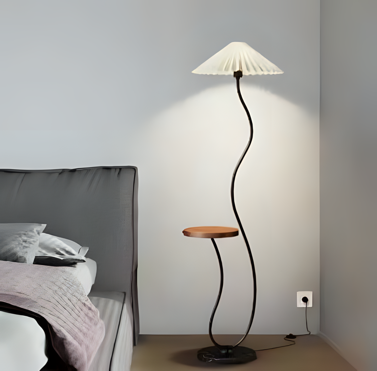 A uniquely designed floor lamp with a wavy black stand, a small wooden side table attached, and a pleated lampshade, placed beside a bed in a modern bedroom.