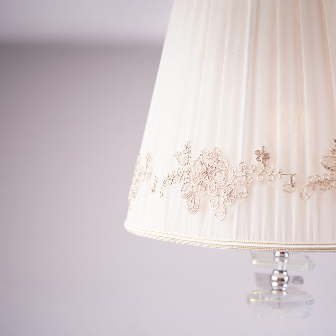 Elegant white table lamp with gold embroidery on the pleated shade, featuring a glass base, placed in a bright setting.