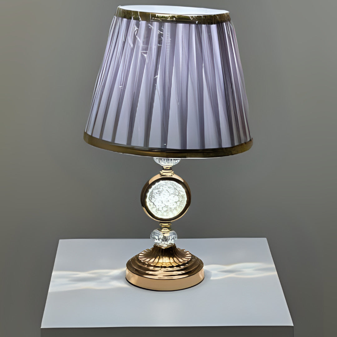 Round gold table lamp with a pleated grey shade with gold trim, featuring a crystal centrepiece, placed on a white table.