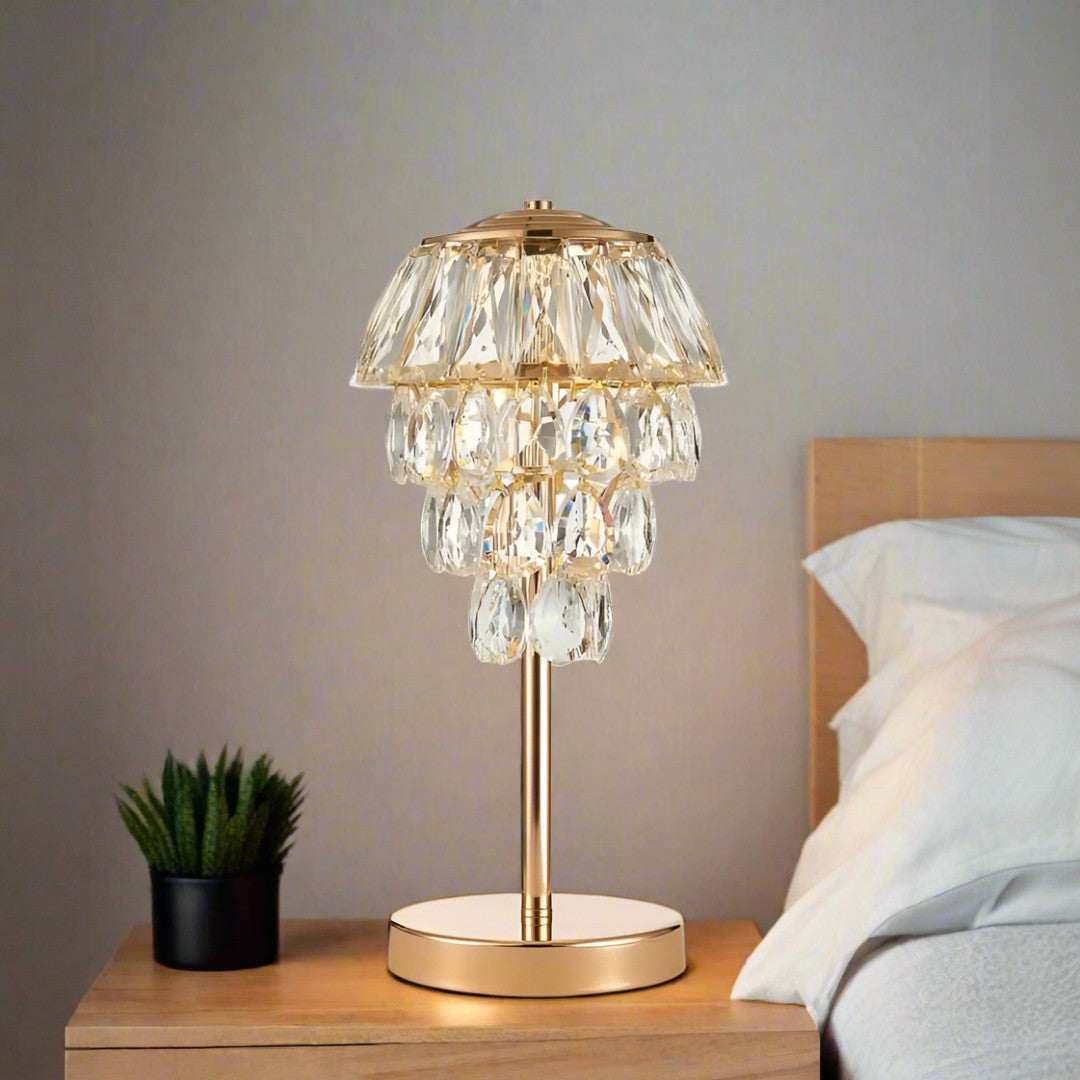 This table lamp features a gold base with a slender stem, topped by cascading crystal pendants arranged in tiers. It was placed on a wooden table behind the bed