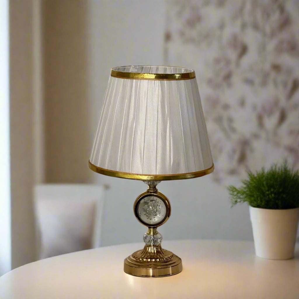 Elegant gold table lamp with a pleated white shade with a gold trim and a crystal accent in the center, placed on a white table.