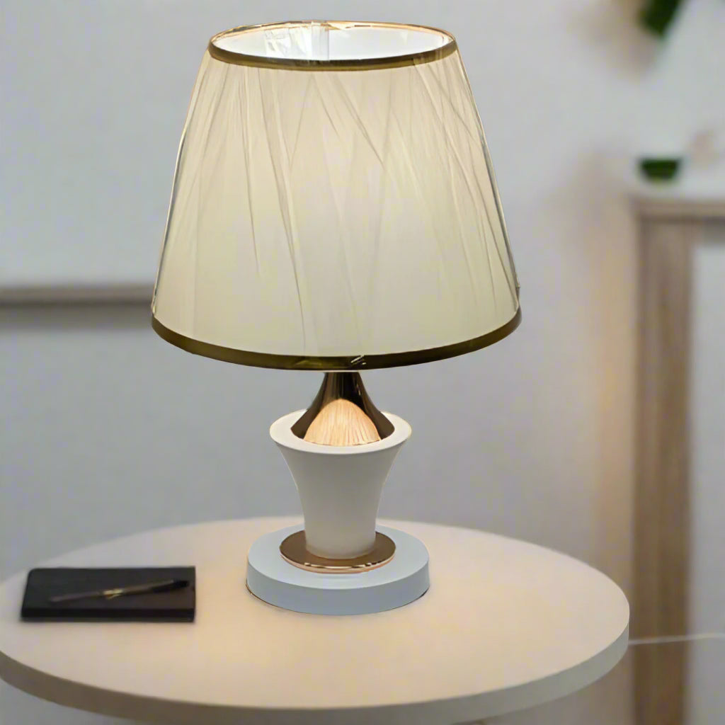 Luxury cream and gold table lamp with a slightly flared pleated lampshade wrapped in protective plastic, placed on a round white table.