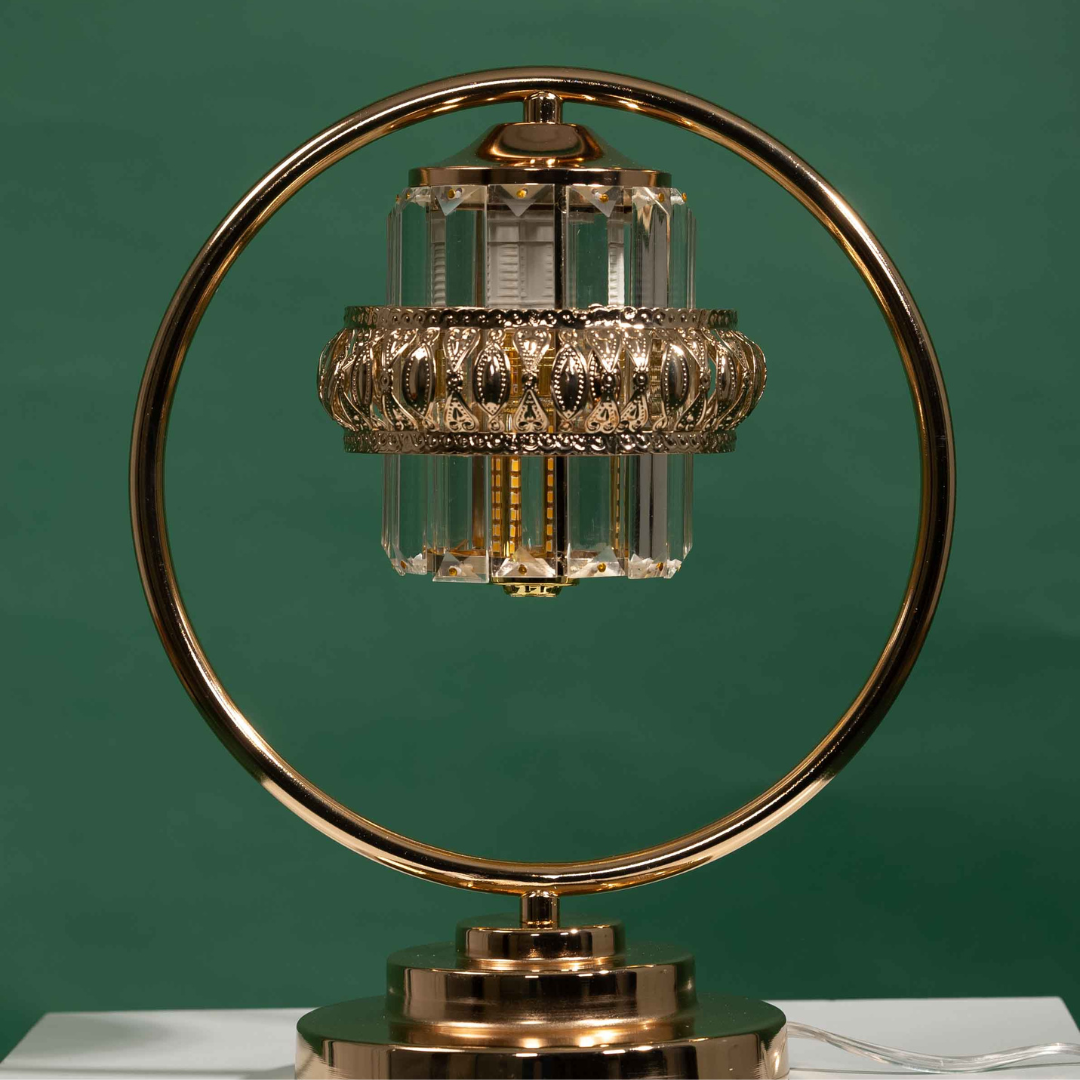 A luxurious table lamp with a circular gold frame and suspended crystal elements, designed to resemble a chandelier. Placed on a round white table and green background.