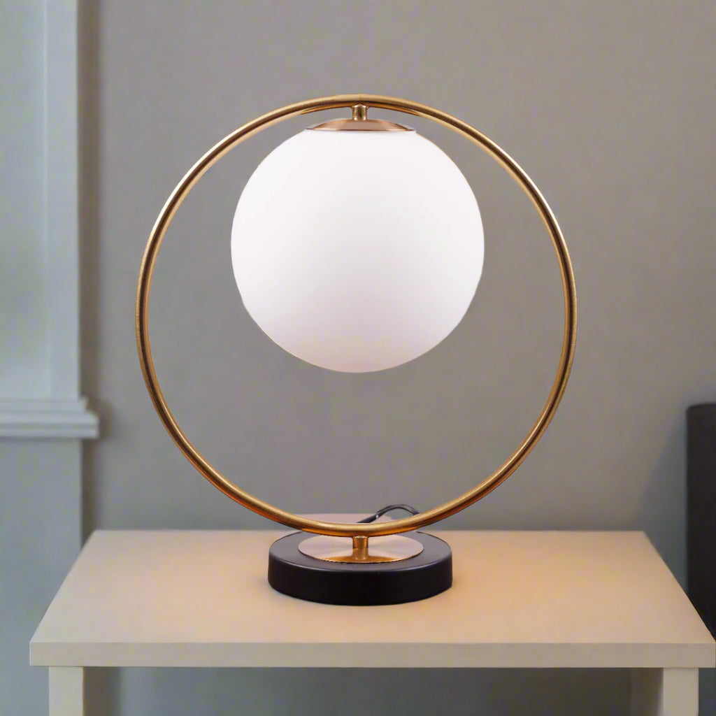 This modern table lamp features a striking circular metal frame that elegantly encases a frosted glass globe, creating a sleek and contemporary aesthetic. Placed on a wooden table.