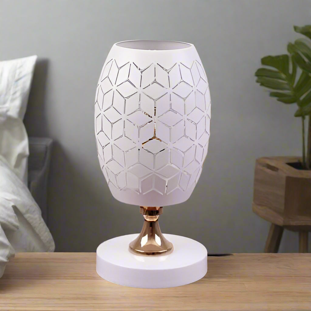 A white modern table lamp with a geometric design, gold accents, and a sleek base sits on a wooden table. It features an LED light with an E27 bulb, casting a warm, stylish glow.