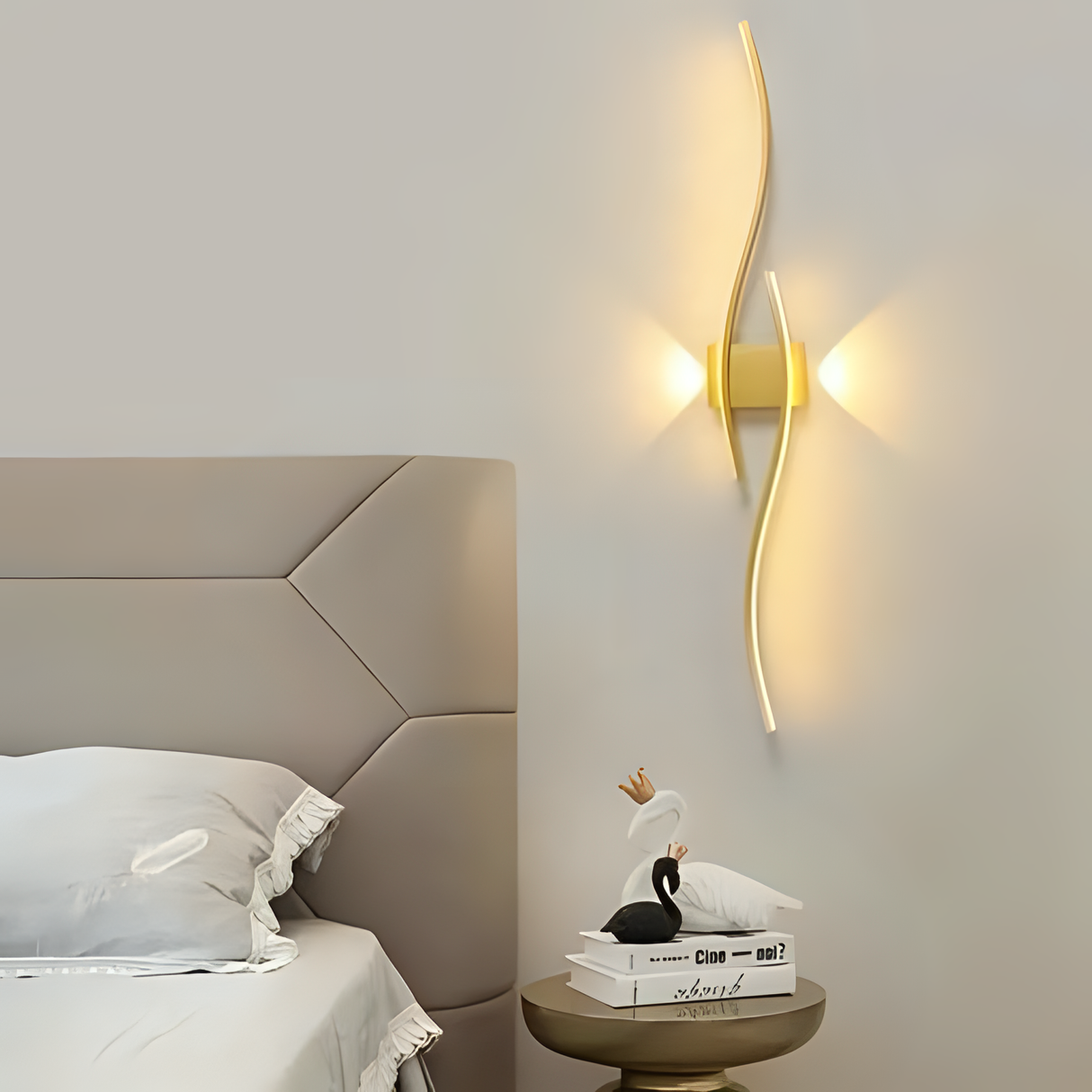 A stylish gold wall sconce with a wavy, elongated LED light design, placed beside a bed with a neutral-toned headboard.