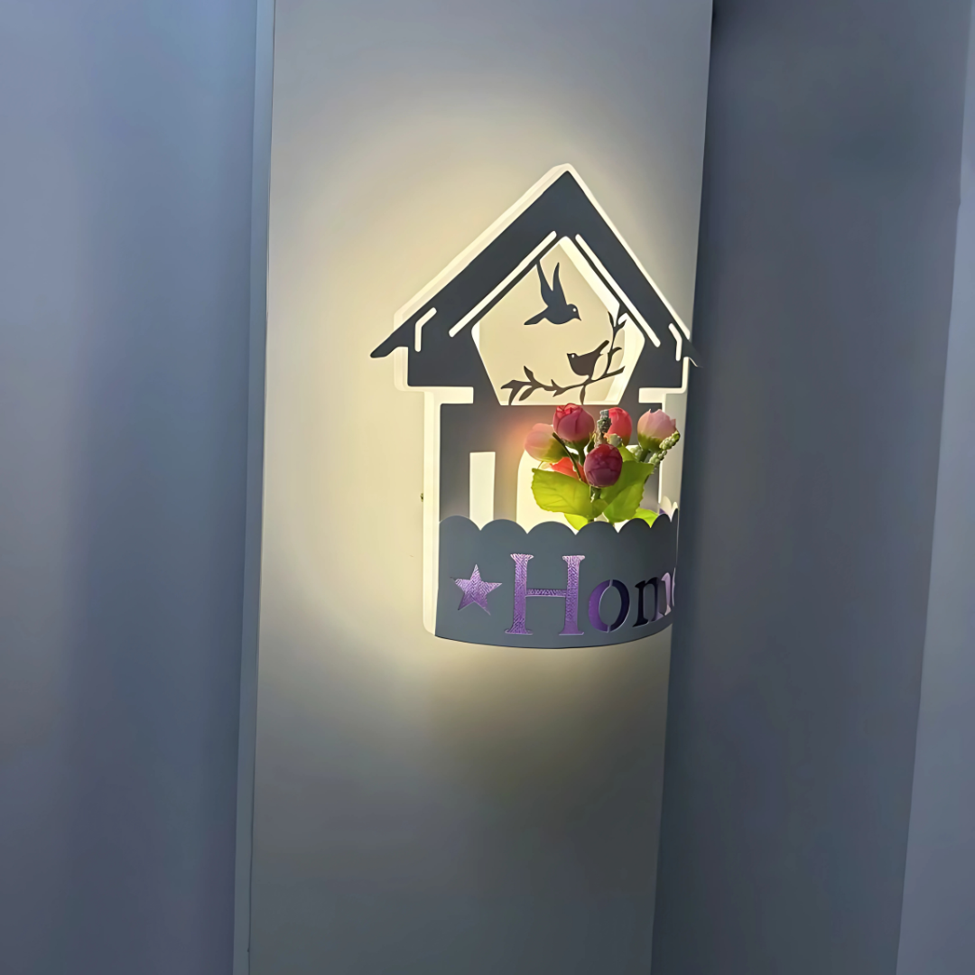 Wall-mounted LED light fixture designed in the shape of a white house , featuring a cork-style built-in compartment holding artificial pink and red tulip flowers. The light has a whimsical, cottage-inspired aesthetic and is mounted on a plain white wall.