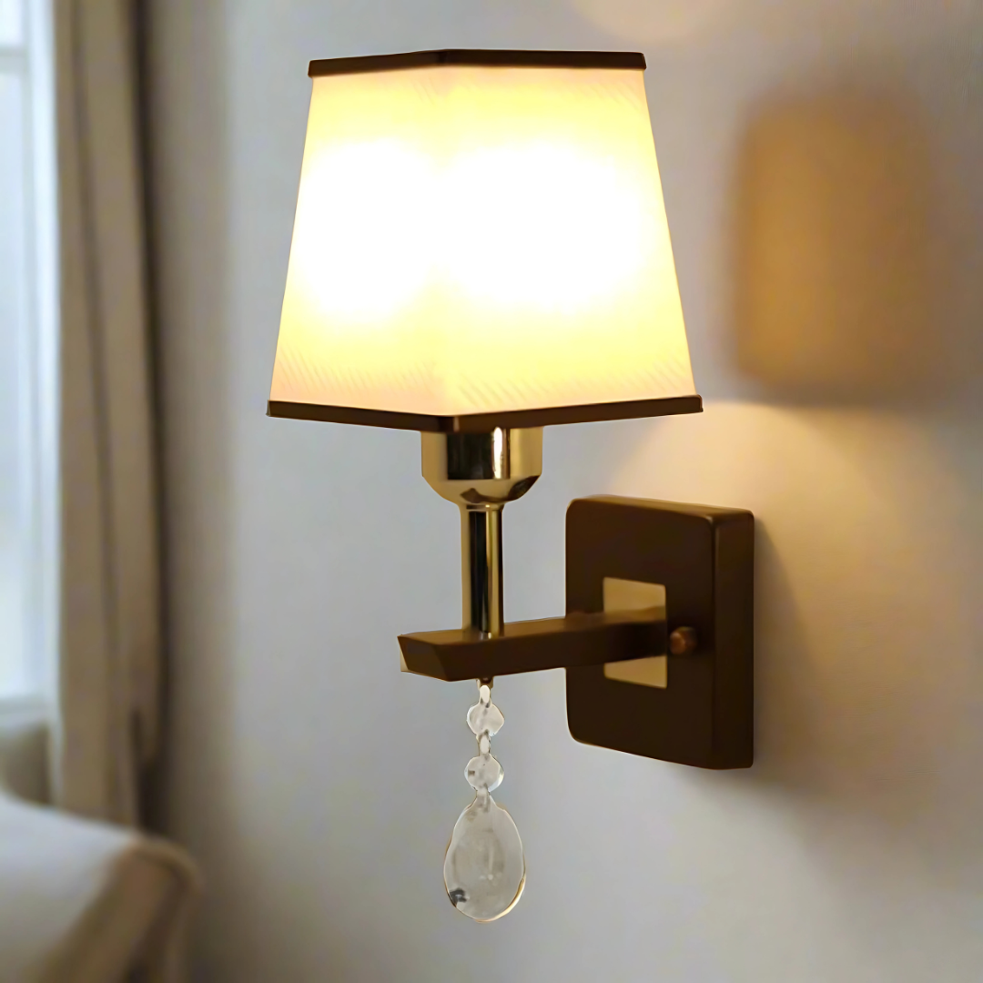 A brown and silver wall light with a rectangular fabric shade and a hanging crystal ornament, adding a touch of elegance turned on, casting warm light.