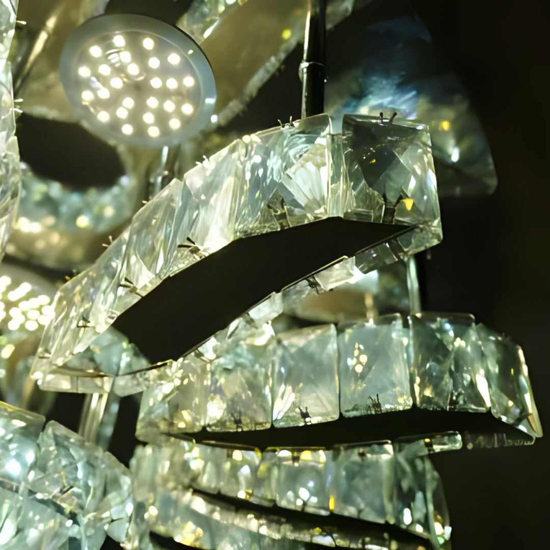 A luxurious crystal chandelier with curved, overlapping rectangular glass segments, suspended from the ceiling in a dark space; it reflects bright white light in multiple directions.