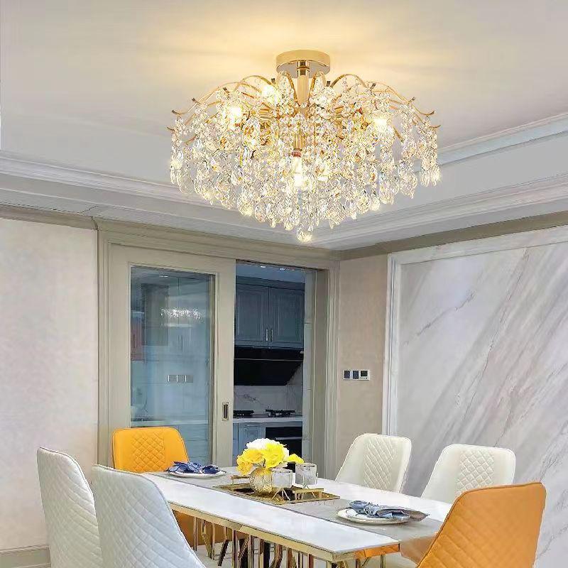 An ornate crystal chandelier with a golden metal base hanging in a modern dining room. It is centered above a rectangular dining table with white and yellow chairs, framed by marble walls.