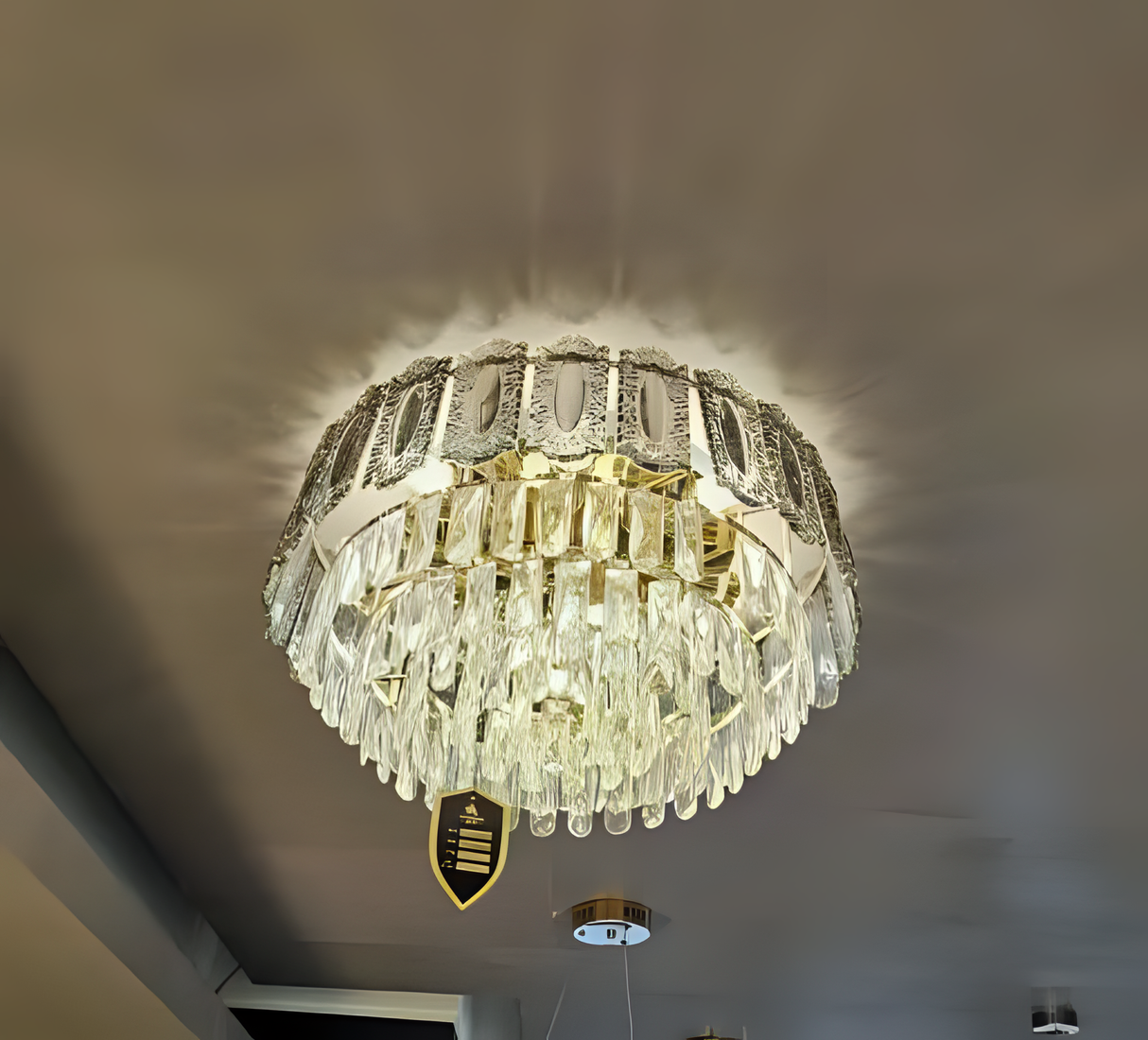 A striking modern chandelier with a drum-like shape made of vertical crystal prisms. The chandelier emits a clear, bright light and is mounted close to the ceiling in a contemporary room with a dark-painted ceiling.