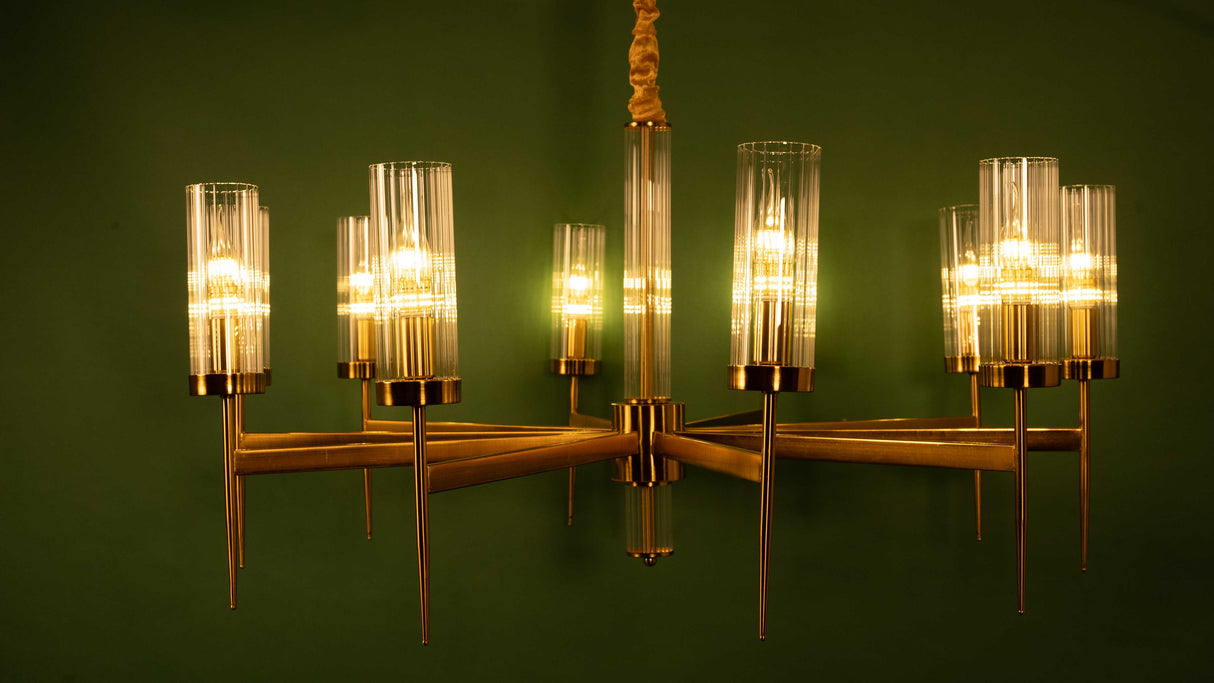 Metal chandelier with symmetrical geometric arms and vertical glass shades against a green wall backdrop.