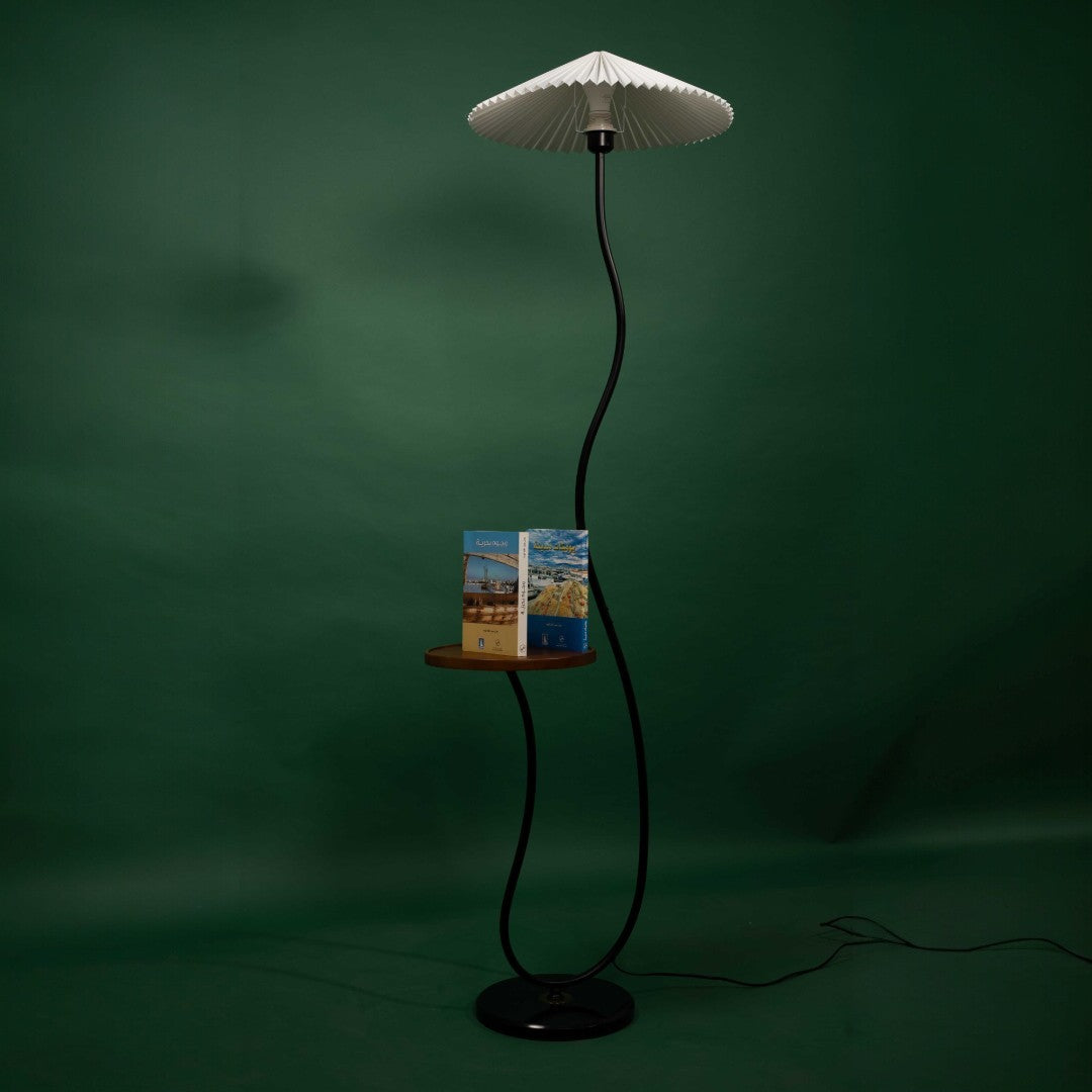 A black floor lamp with a wavy stand, a pleated white lampshade, and a small wooden shelf holding books, photographed against a green background.