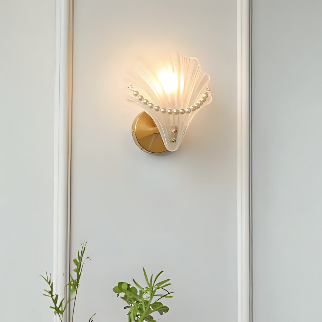 A decorative seashell-shaped wall sconce with a gold circular base and a sculpted white glass shell adorned with pearl-like accents, emitting a warm glow.