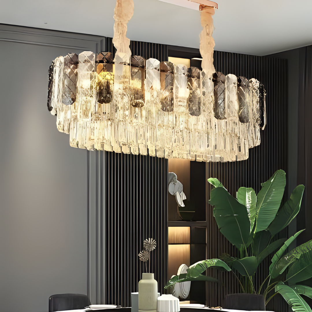 Elegant oval crystal chandelier with layered, clear and smoky glass prisms suspended from fabric-wrapped supports, casting a warm and luxurious glow in a modern dining room setting with dark paneling and lush green plants.