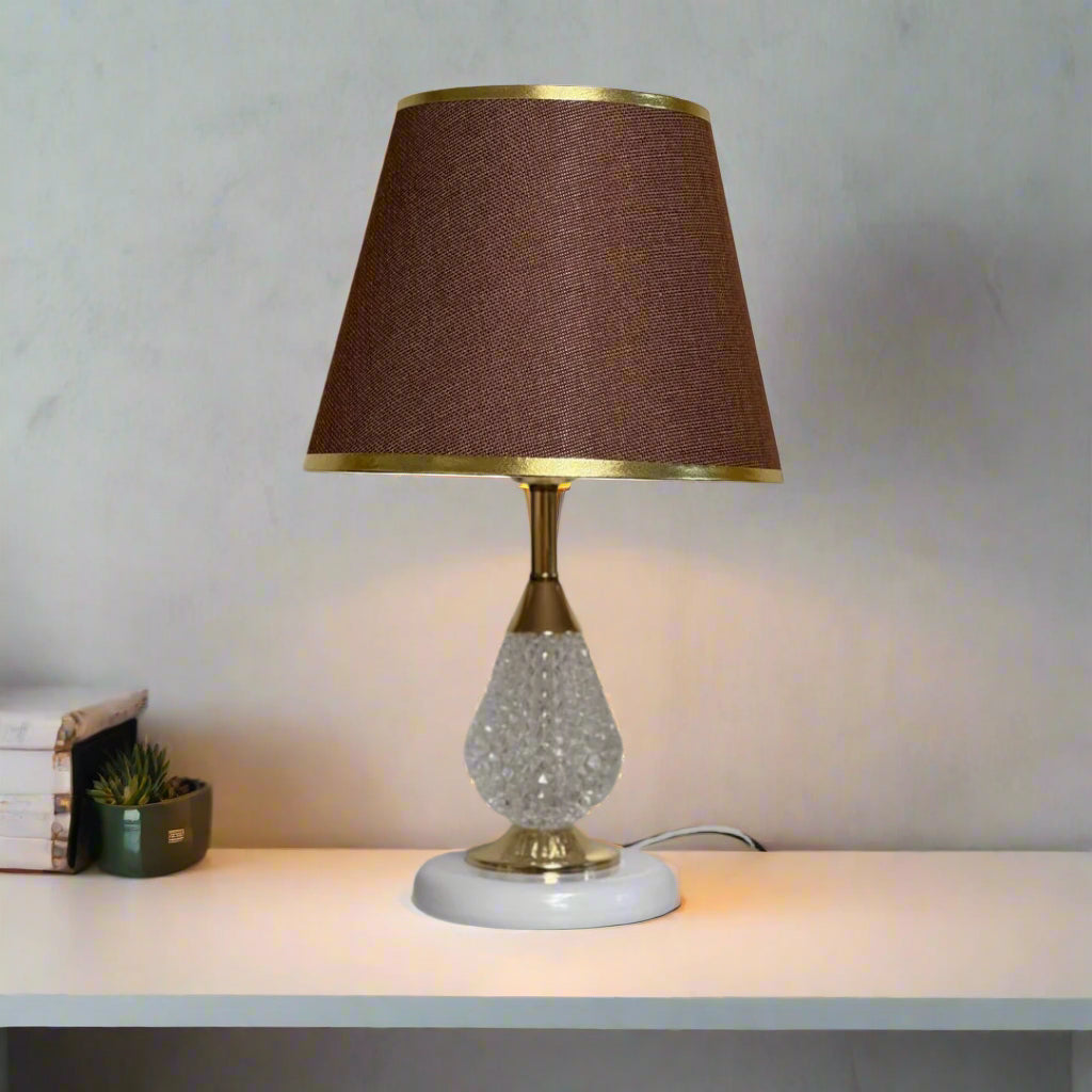 A table lamp with a crystal-textured glass base, gold detailing, and a brown fabric lampshade with gold trim, set against a plain light-colored wall.
