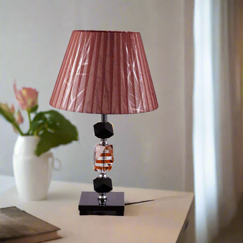 An elegant brown table lamp featuring a classic white lampshade and a sleek black-and-transparent glass base, placed on a wooden table. Equipped with an E27 LED light, it radiates a warm and sophisticated glow, enhancing the ambiance with a touch of refinement.