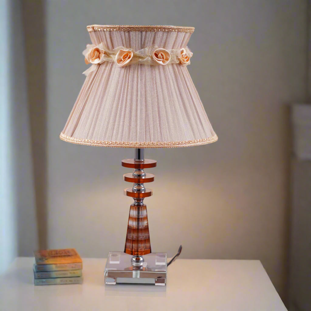 Elegant table lamp with a beige pleated lampshade, adorned with delicate fabric roses and gold trim. The base is crafted from a combination of transparent and amber-colored glass with a geometric design, supported by a sturdy glass platform. Equipped with an E27 LED light, it emits a warm and sophisticated glow. Placed on a white wooden table.
