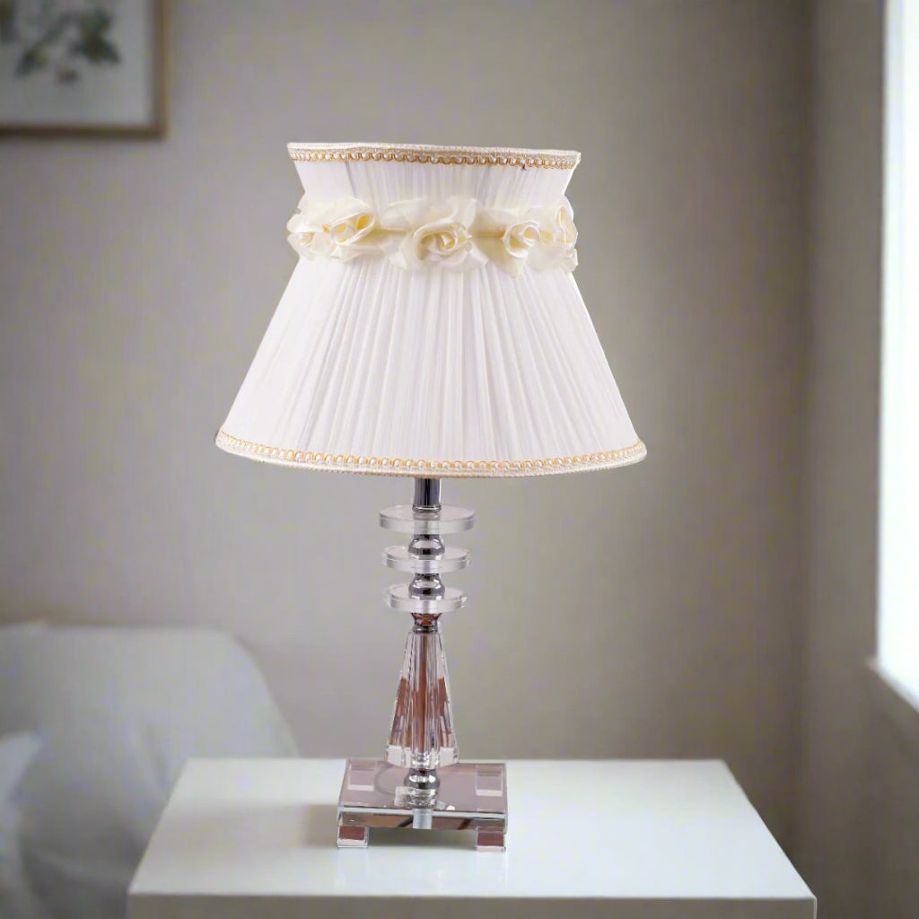 Elegant table lamp with a white pleated lampshade, adorned with delicate fabric roses and gold trim. The base is crafted from a combination of transparent and amber-colored glass with a geometric design, supported by a sturdy glass platform. Equipped with an E27 LED light, it emits a warm and sophisticated glow. Placed on a white table.