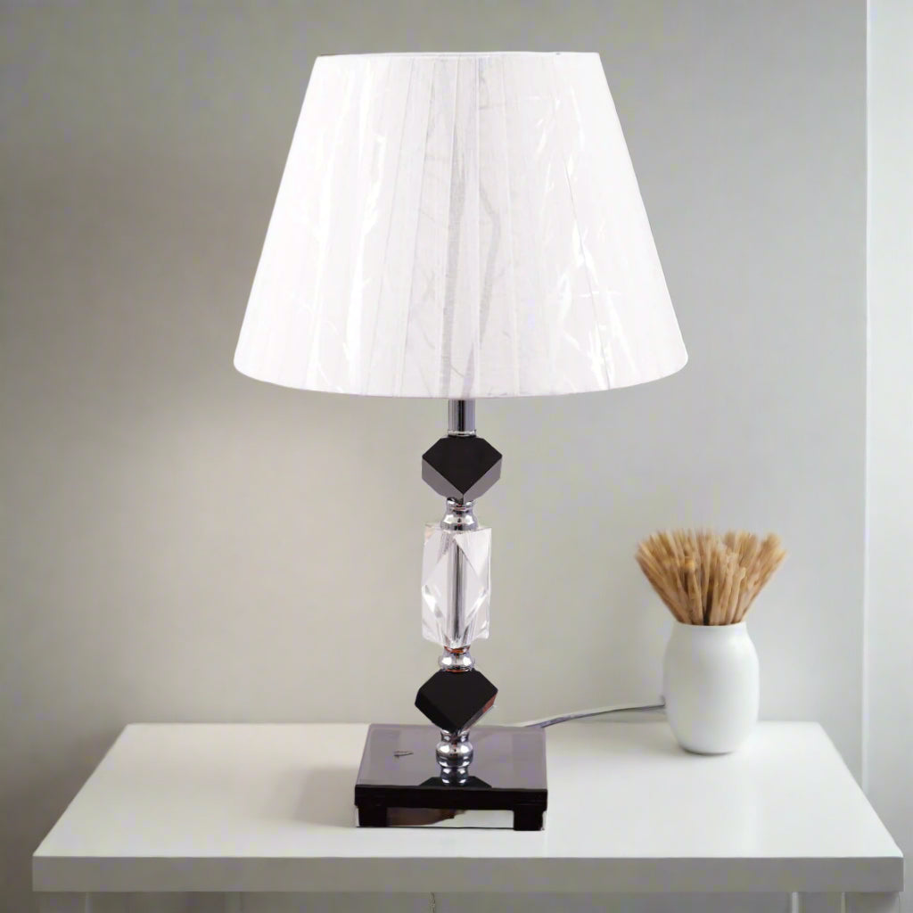 An elegant table lamp featuring a classic white lampshade and a sleek black-and-transparent glass base, placed on a wooden table. Equipped with an E27 LED light, it radiates a warm and sophisticated glow, enhancing the ambiance with a touch of refinement.