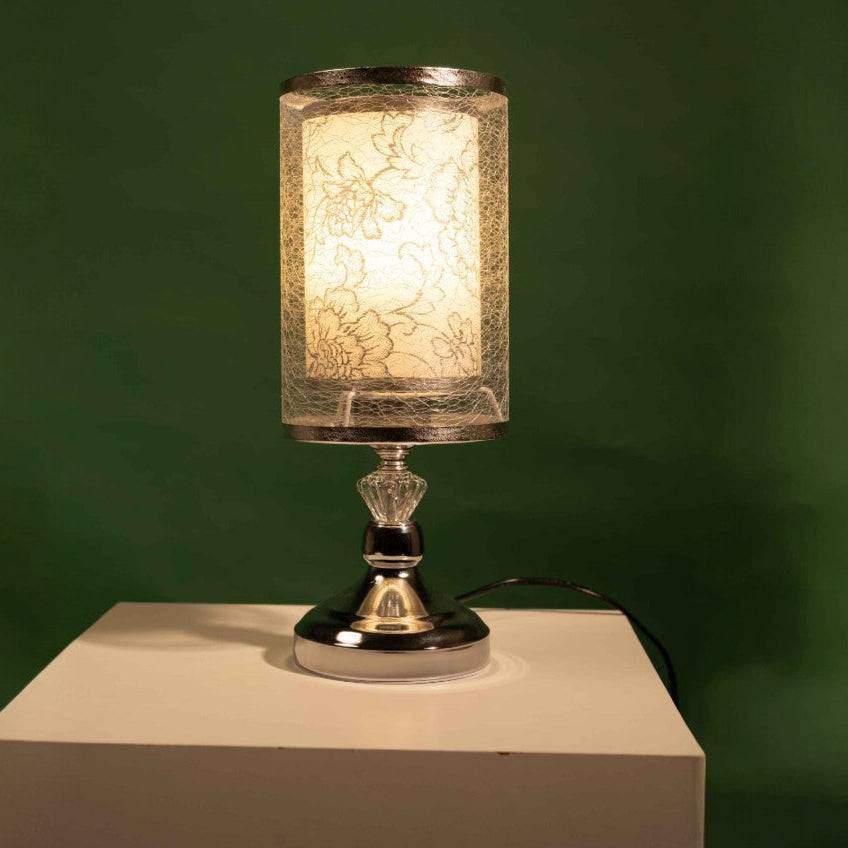 Modern silver table lamp with a cylindrical transparent lampshade featuring delicate floral patterns, placed on a white table and green background.