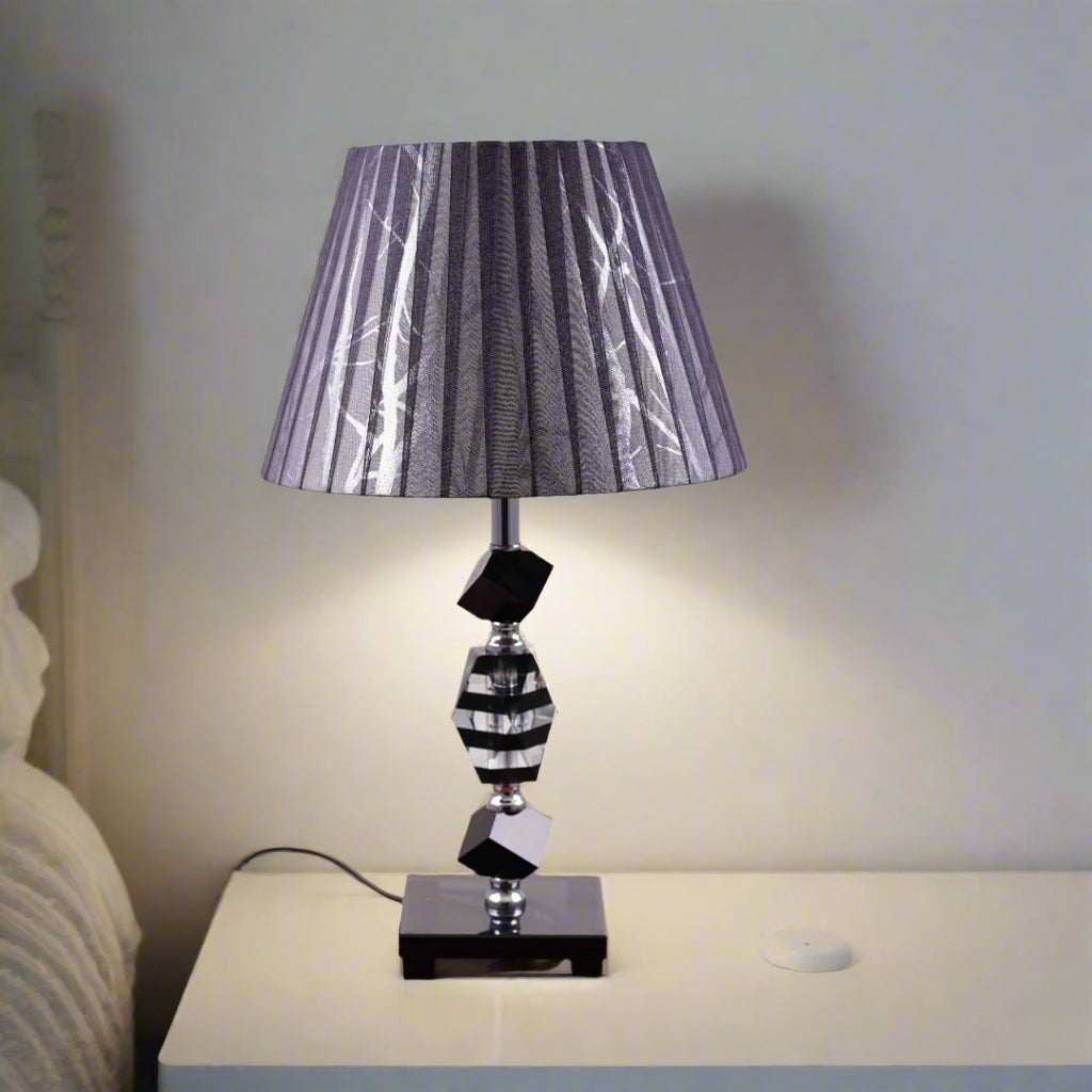 An elegant black table lamp featuring a classic white lampshade and a sleek black-and-transparent glass base, placed on a wooden table. Equipped with an E27 LED light, it radiates a warm and sophisticated glow, enhancing the ambiance with a touch of refinement.