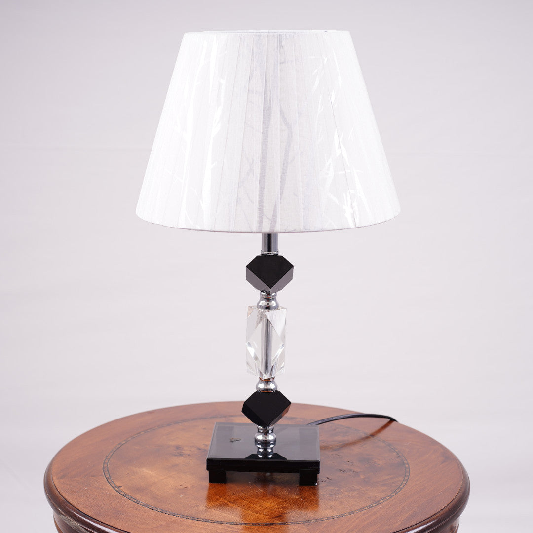 An elegant white table lamp featuring a classic white lampshade and a sleek black-and-transparent glass base, placed on a wooden table. Equipped with an E27 LED light, it radiates a warm and sophisticated glow, enhancing the ambiance with a touch of refinement.