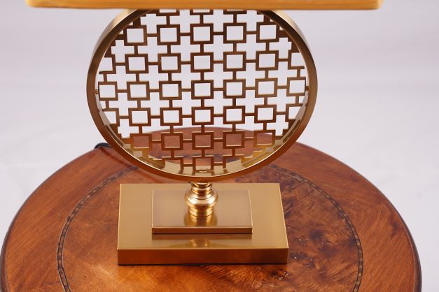 A modern table lamp featuring a gold metal geometric base with intricate latticework, placed on a round wooden table.